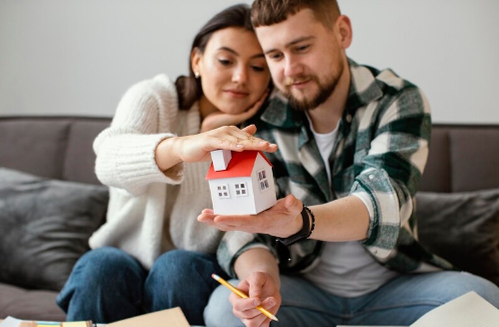 A young couple sitting on a couch, holding a small model house together while planning finances, symbolizing joint investment decisions and accessible opportunities through real estate crowdfunding.