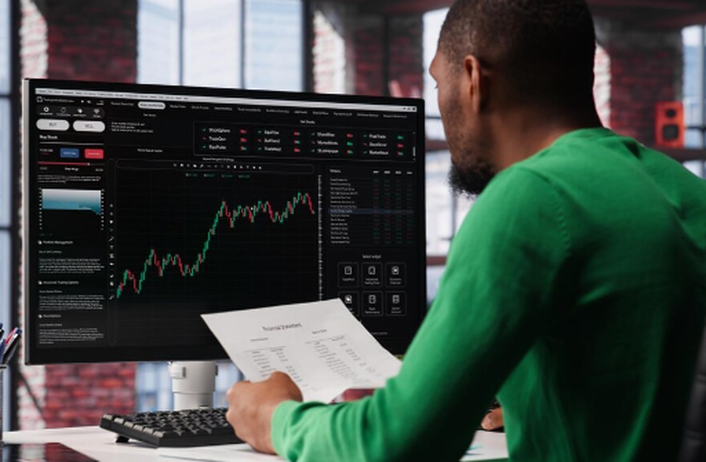 A trader analysing stock market charts on a computer screen while holding financial reports, representing strategies and decision-making in day trading.