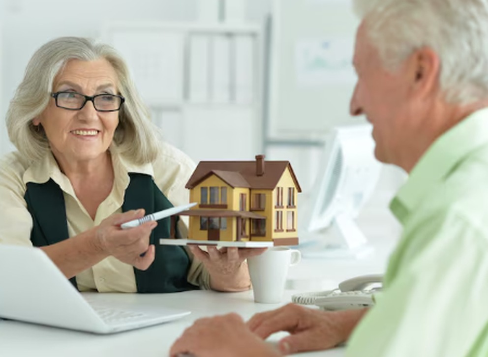 An elderly couple discussing property investment with a miniature house model on the desk, symbolising guidance and opportunities in the estate market.